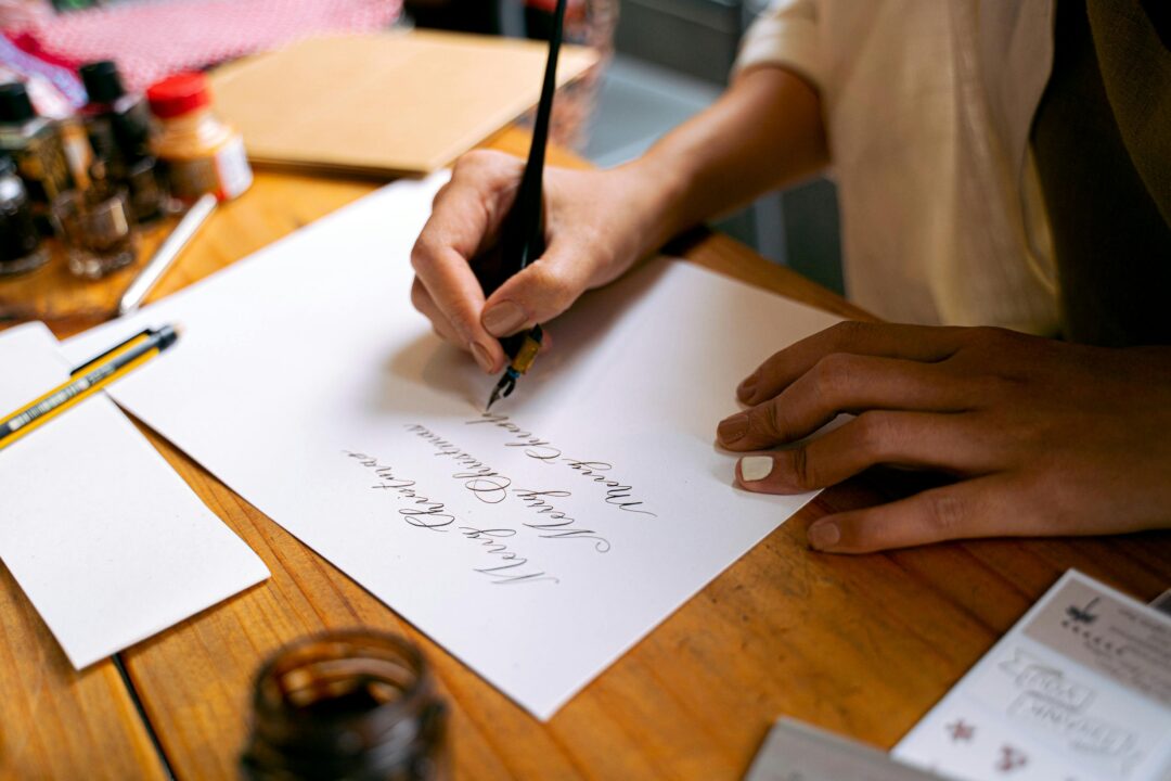 Person handwriting a personal note inside a Christmas card on a wooden desk, festive minimal styling with pen and evergreen sprig, intimate close-up n