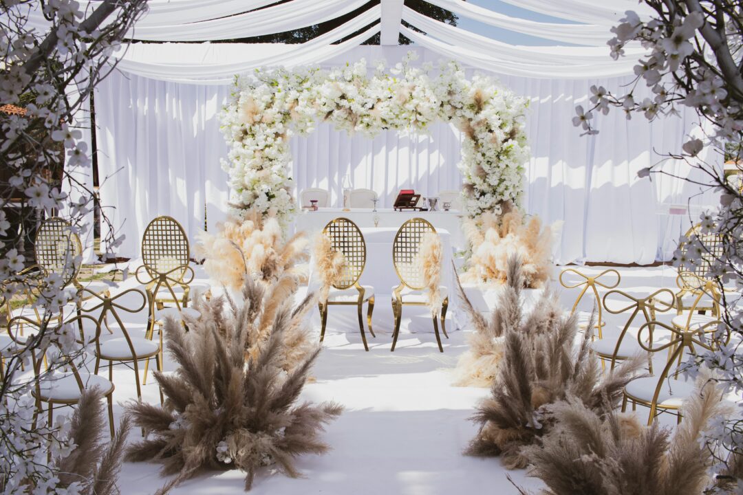 Emerald and gold wedding ceremony arch with cascading tropical greenery, white garden roses, gold geometric frame accents, and hanging gold botanical 