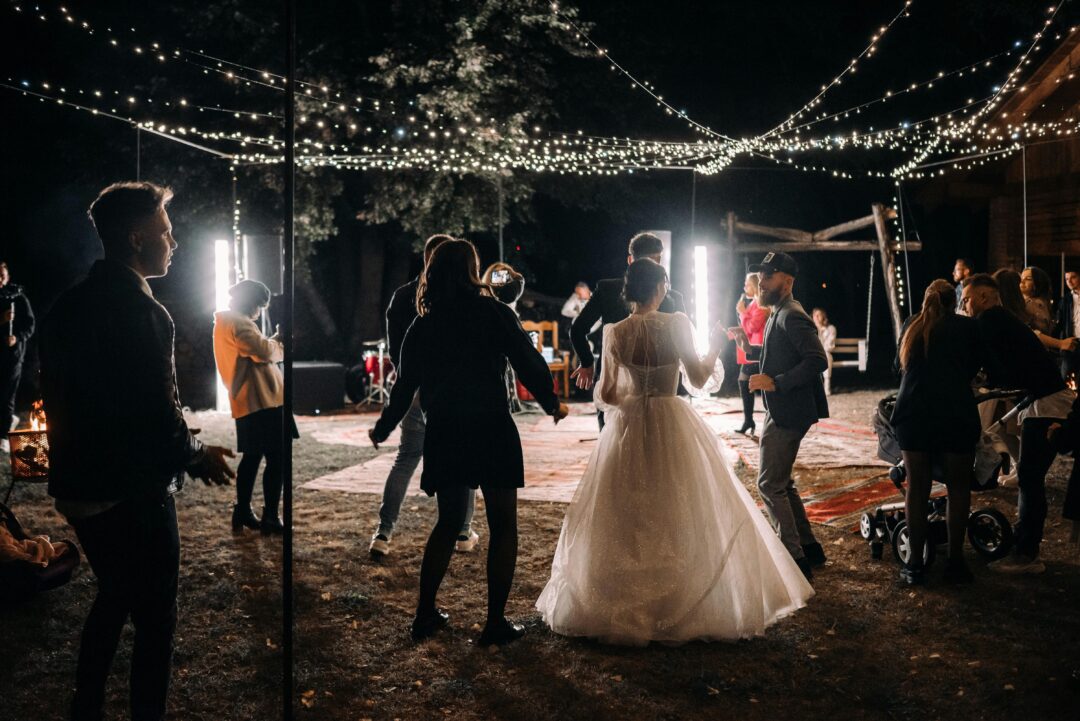 wedding DJ at the decks checking a tablet, warm reception lighting, visible dance floor and guests in soft focus behind