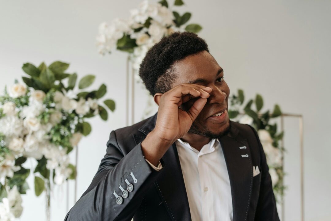 groom wiping away laughter-tears during ceremony while bride grins, wedding party smiling in background, documentary-style candid shot
