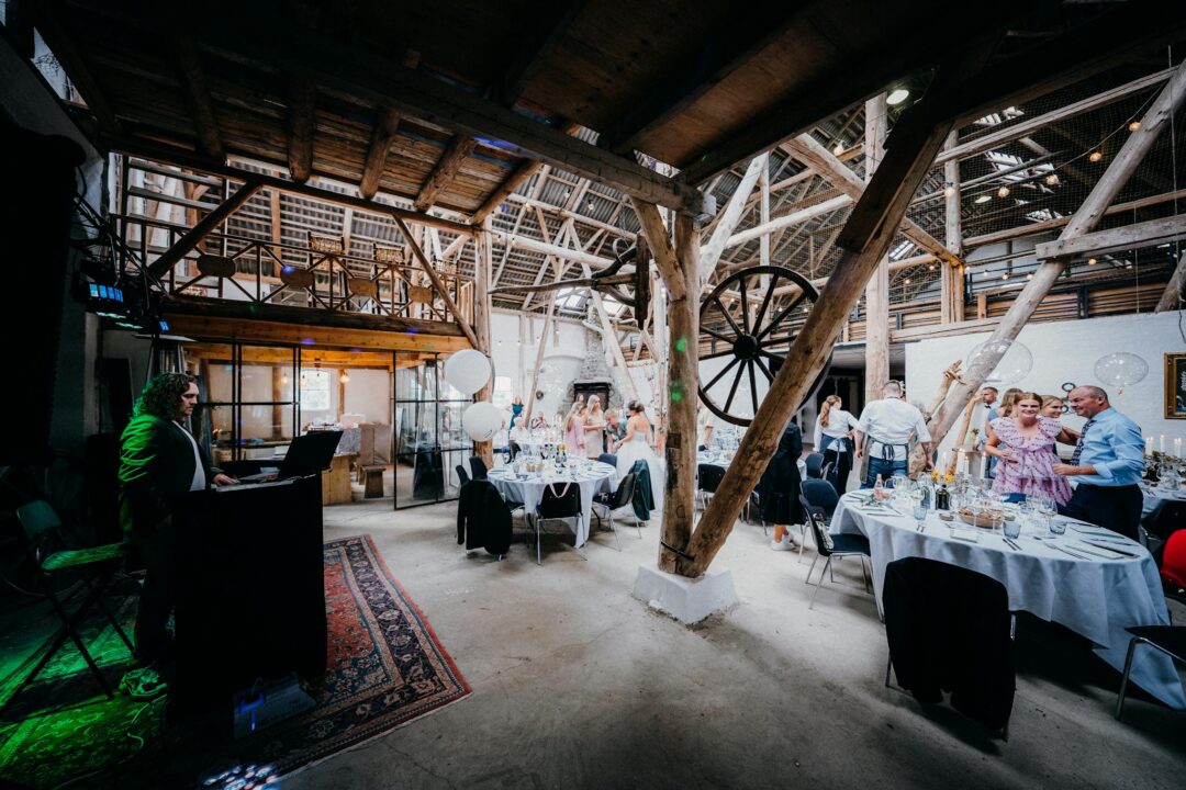 rustic barn wedding reception with DJ booth at left, guests gathered at candlelit tables under exposed timber beams