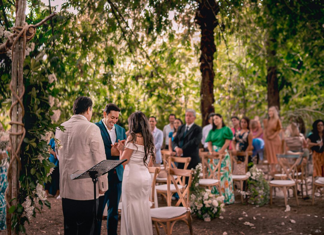 couple standing at ceremony altar smiling at each other while guests laugh, wide shot showing decorated outdoor venue with string lights