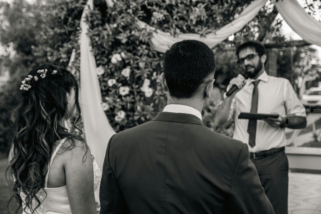 officiant standing with couple at outdoor ceremony, holding ceremony script, couple facing each other, guests seated in rows behind, garden or natural