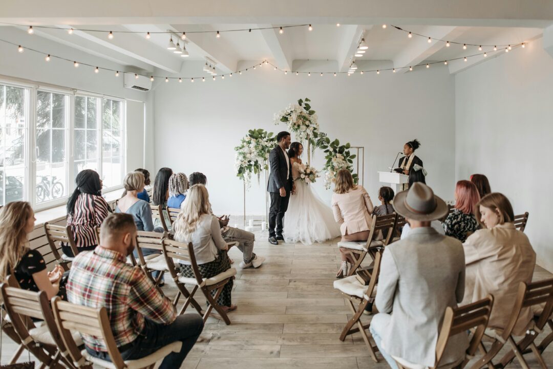 wedding ceremony venue from guest perspective, couple standing at altar with officiant, lush greenery or floral arrangements framing the scene, bright