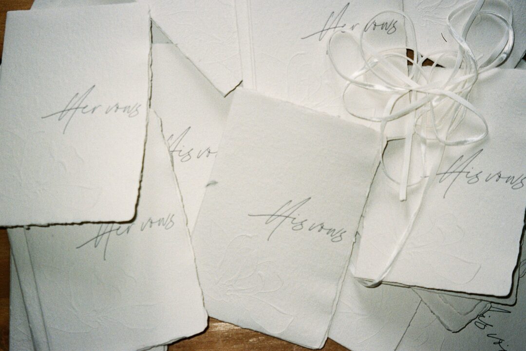 elegant vow cards printed on thick cotton paper resting on ceremony program, letterpress texture visible, beside fresh white peonies, flat lay styling