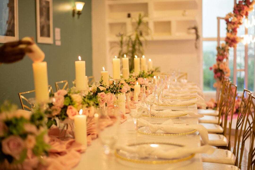 Intimate wedding reception dinner in progress at a long wooden table, guests passing platters family-style, warm candlelight and greenery ru