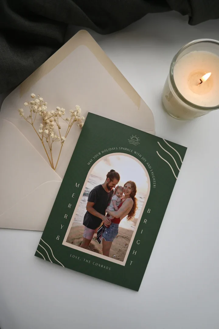 Dark green Christmas card with a round window showing a family beach photo, gold metallic text, dried flowers, and a candle on a light surface.