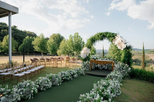 wide-angle outdoor wedding ceremony aisle lined with white florals and greenery, soft natural light, romantic garden or barn setting