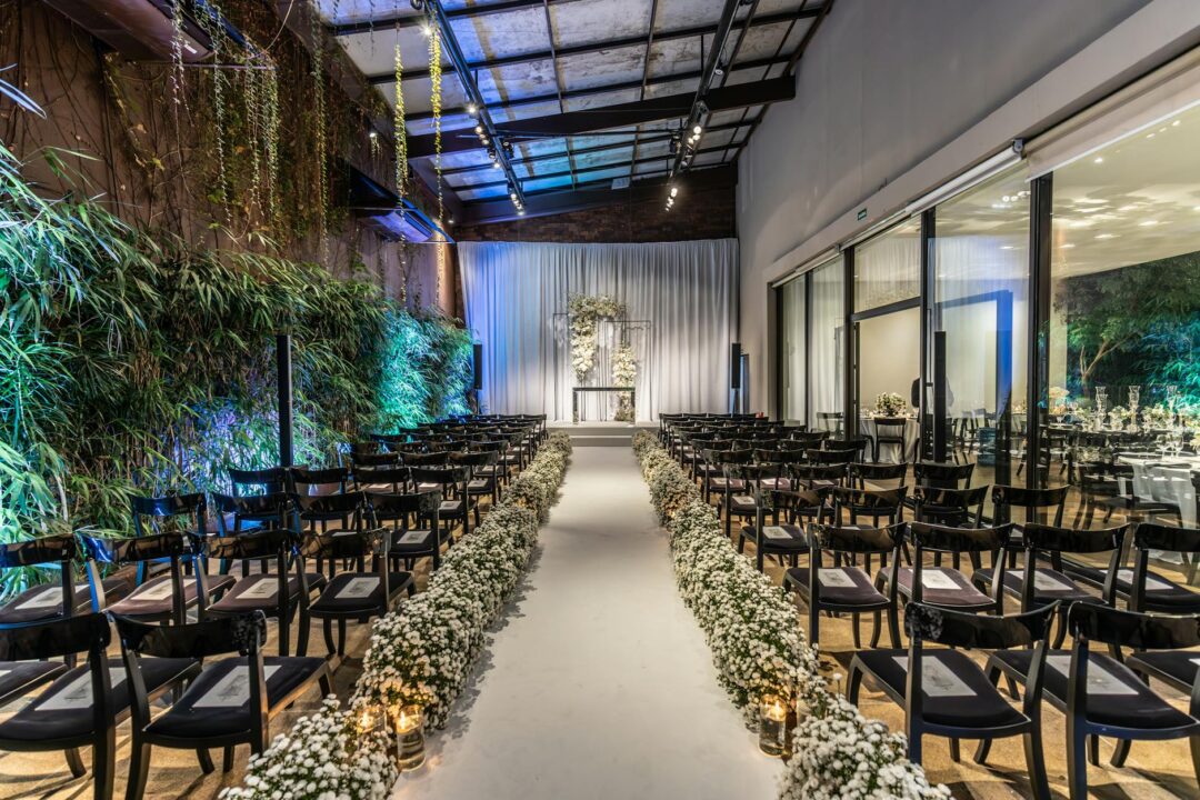 indoor wedding ceremony with white floral arch in a ballroom, chandeliers above, guests seated in rows
