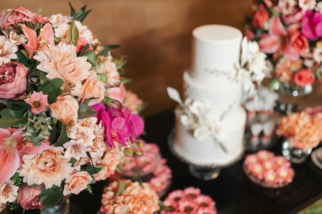 A three-tiered white wedding cake surrounded by vibrant pink and peach flower bouquets.