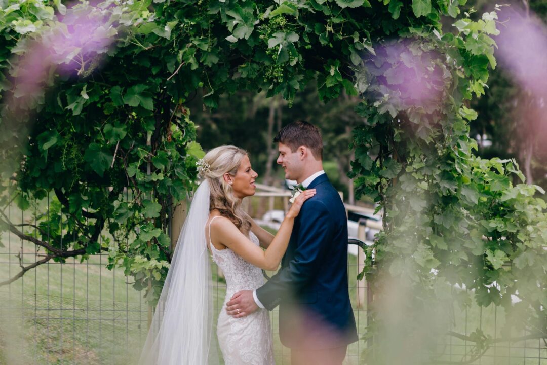 Australian outdoor wedding ceremony scene, celebrant speaking at a floral arch with guests seated in rows on grass, golden hour light
