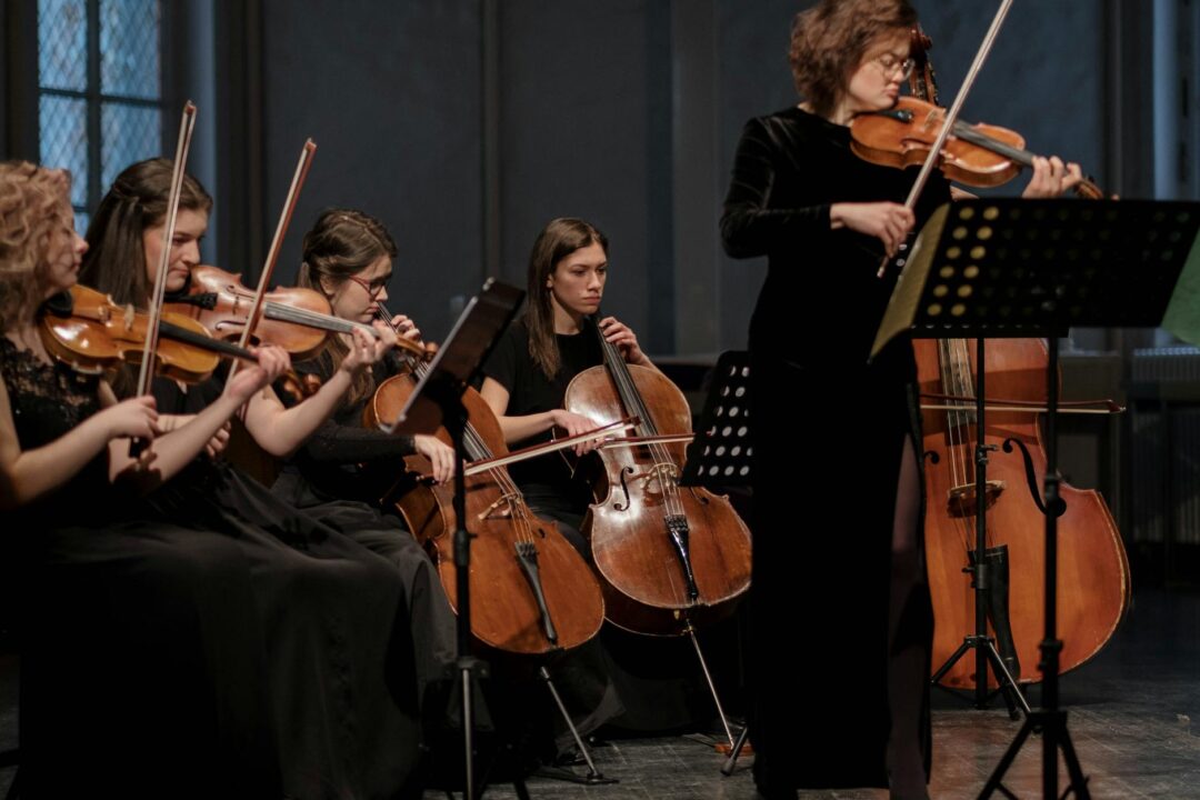 A group of women musicians performing classical music with violins and cellos in an orchestral setting.