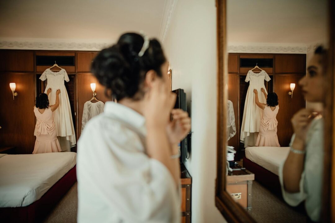 bride and groom getting ready at a bright dressing room mirror, bridal party in background, morning light, editorial wedding photography style