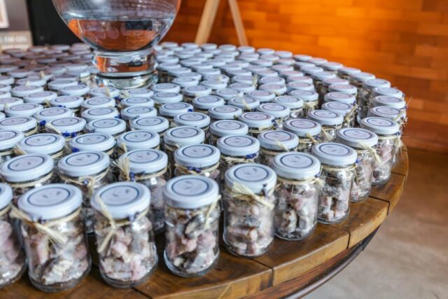 styled wedding favor display with small glass jars and kraft boxes lined up on a rustic wooden table, each tied with twine and handwritten t