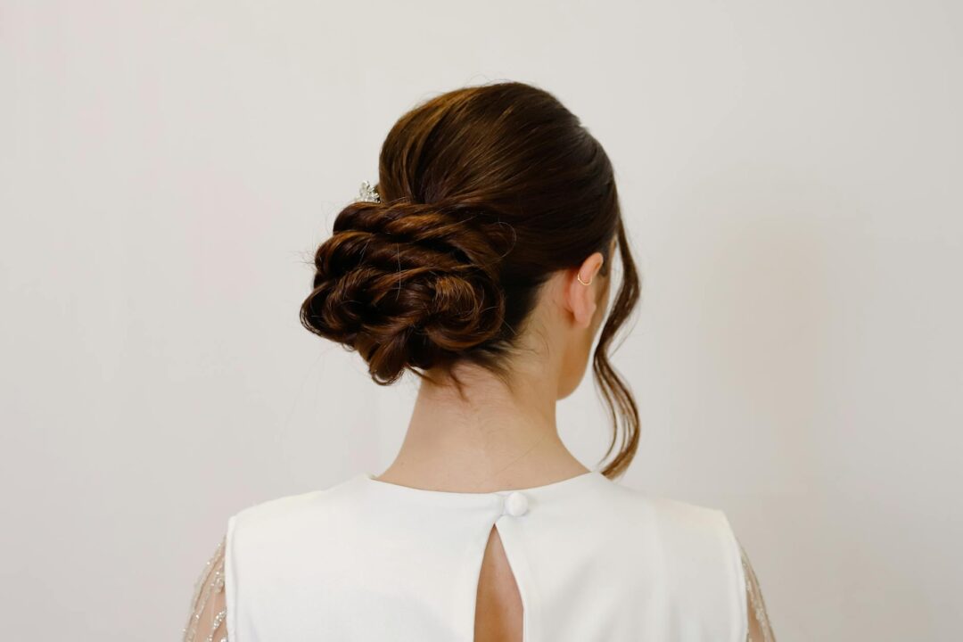 Bride with a soft low bun at the nape, wearing a fitted lace wedding gown with an open back, loose face-framing tendrils visible, photograph