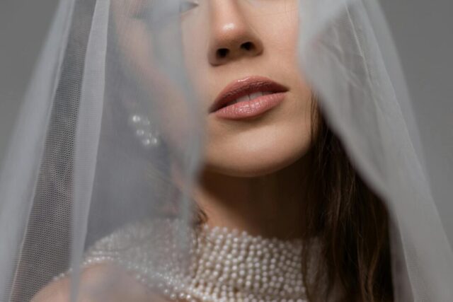 close-up bridal portrait of bride with a romantic low chignon adorned with a cluster of pearl-head pins and delicate face-framing tendrils, wearing a lace off-shoulder wedding gown, warm natural studi