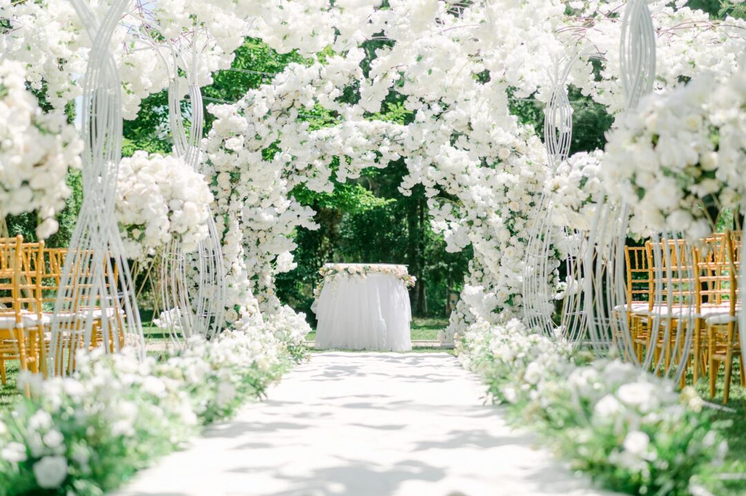 Wedding ceremony outdoor aisle with floral arch, couple exchanging vows, guests seated on either side in sunlit garden venue