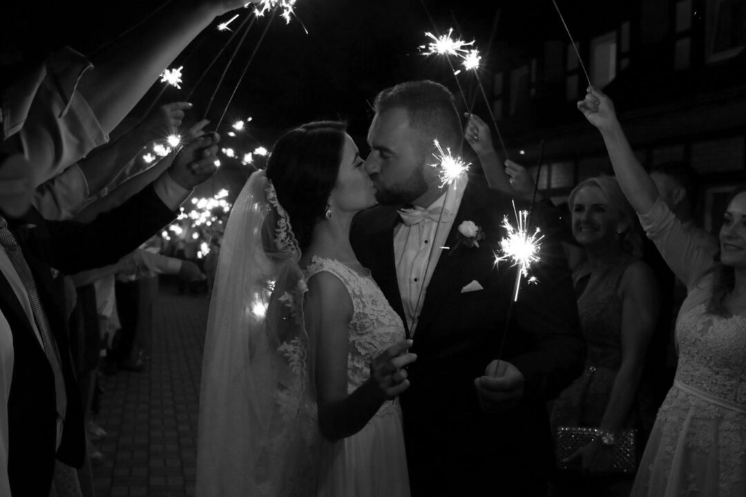 Wedding couple sparkler exit at night, guests lining the path on both sides, warm glowing light trails framing the couple