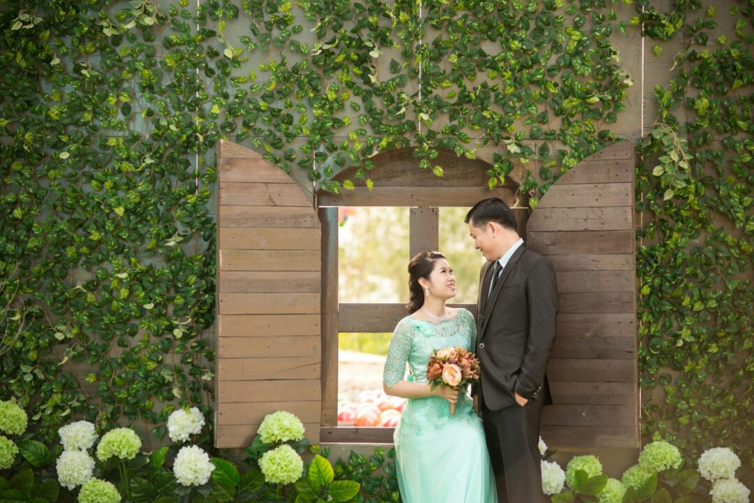 Outdoor garden wedding couple portrait framed through floral arch, dappled natural light, lush greenery background