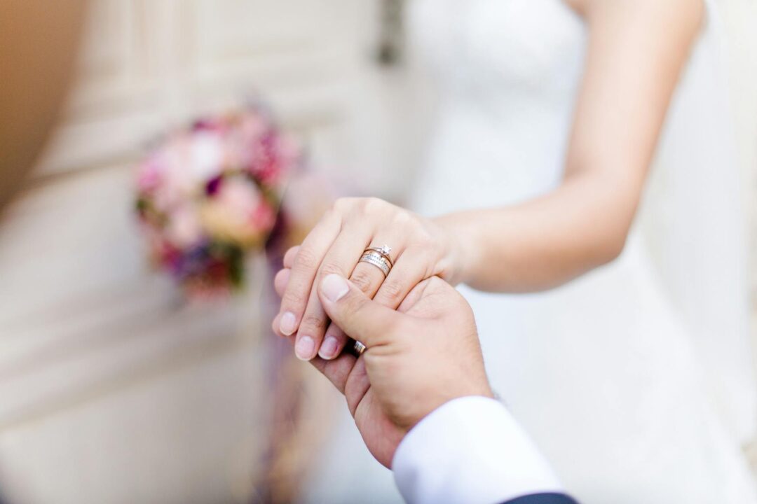 Close-up of wedding couple holding hands with rings visible, bouquet resting against the bride's wrist in warm afternoon light