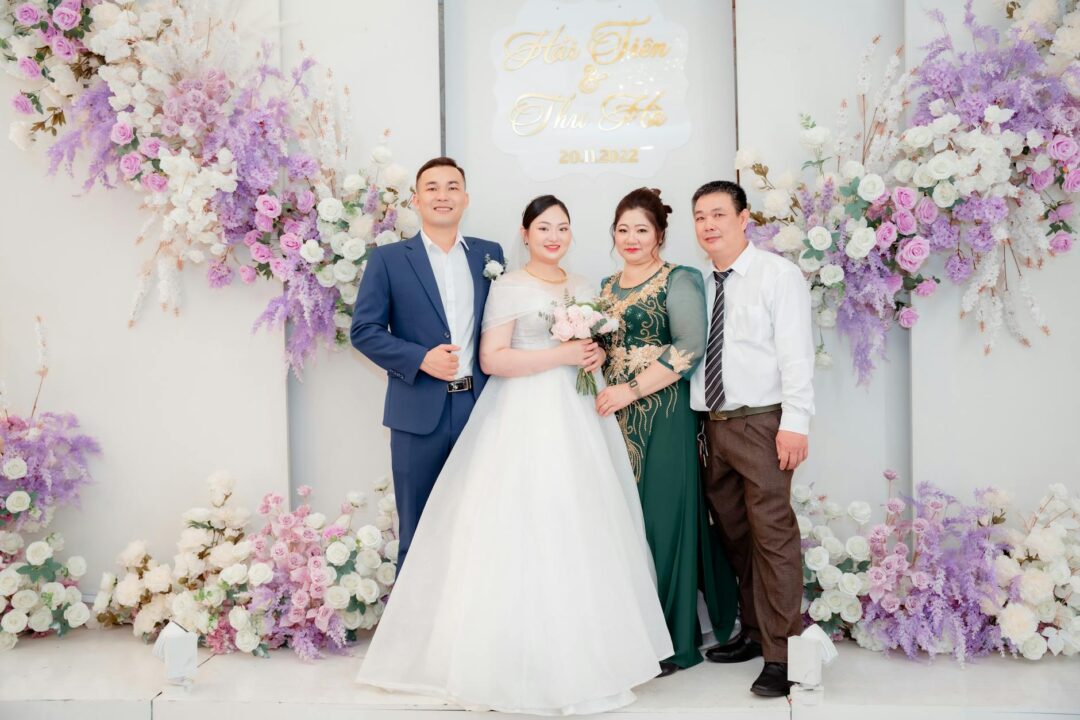 Wedding couple posing with both sets of parents outdoors at a garden venue, everyone smiling naturally and looking at the camera