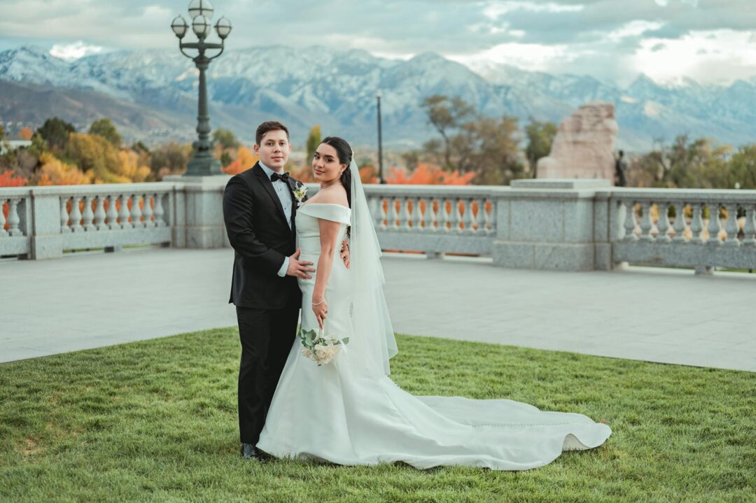 Elegant couple standing together outdoors on their wedding day, groom's hand on bride's waist, both turned slightly toward each other in nat