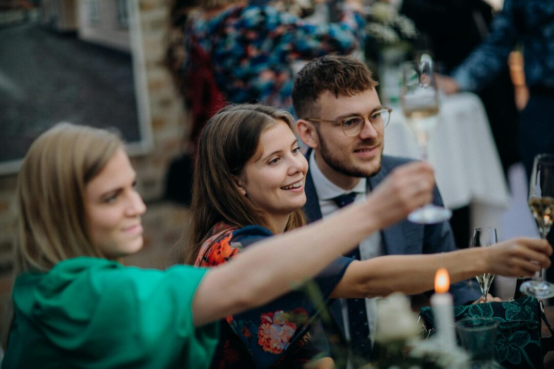 Best man in a navy suit raising a champagne flute and speaking confidently at a beautifully set wedding reception table with white floral ar