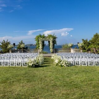 Elegant outdoor wedding ceremony setup with lush floral arch, warm golden light, and cream-colored chairs arranged in garden setting