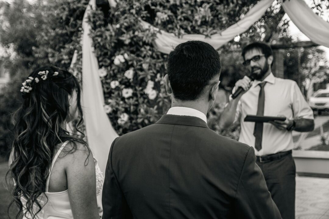 A couple stands facing a speaker during their outdoor wedding ceremony.