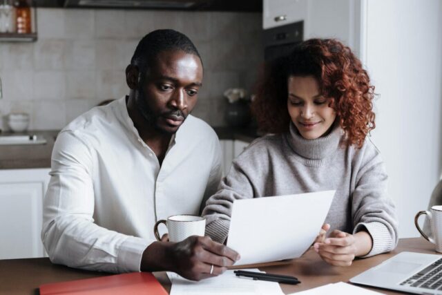 Couple sitting at a kitchen table together reviewing a wedding budget notebook, warm natural light, coffee cups nearby, relaxed planning moo