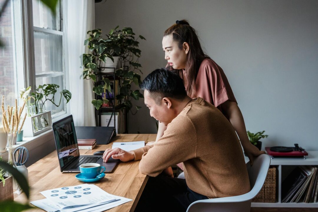 Couple looking at a laptop together at a desk with wedding planning notes and stationery samples spread around them, warm indoor lighting