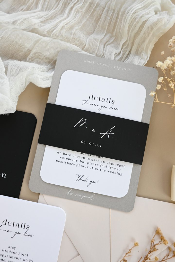Paperlust invitation, Beautifully styled wedding stationery suite flatlay featuring a grey details card with black belly band showing 'M & A' monogram and 05.09.24 date. Elegant neutral tones with soft fabric and dried floral props create a premium aesthetic.
