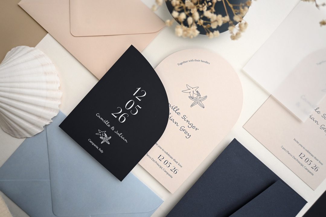 Paperlust invitation, Beautifully styled flat lay of a coastal-themed wedding suite featuring arch-shaped cards in navy and blush with starfish motifs