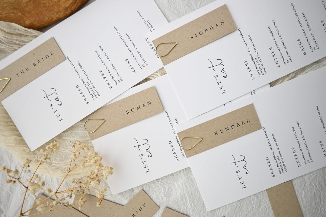 Paperlust invitation, Beautifully styled flatlay of wedding menus with kraft paper name card bands and gold heart clips. Soft