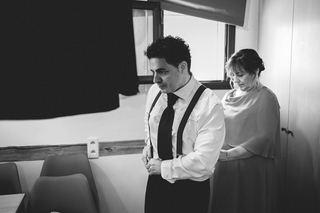 A younger man in formal wedding attire (white shirt, black tie and vest) stands beside an older woman in elegant formal wear. She has her hand on his arm in a warm, affectionate ge