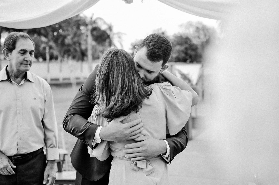 A younger man in formal attire embraces an older woman in a warm hug at an outdoor wedding reception under a tent, with guests visible in the background.