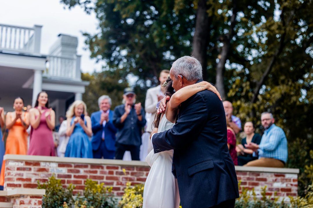 An older man in a dark navy suit embraces a woman in a white wedding dress during an outdoor wedding reception. Wedding guests in formal attire watch from behind a brick wall, appl