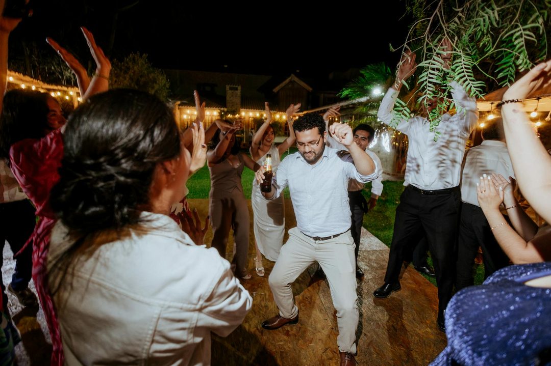 Multiple guests dancing together on an outdoor wedding reception dance floor at night, with string lights and greenery visible in the background