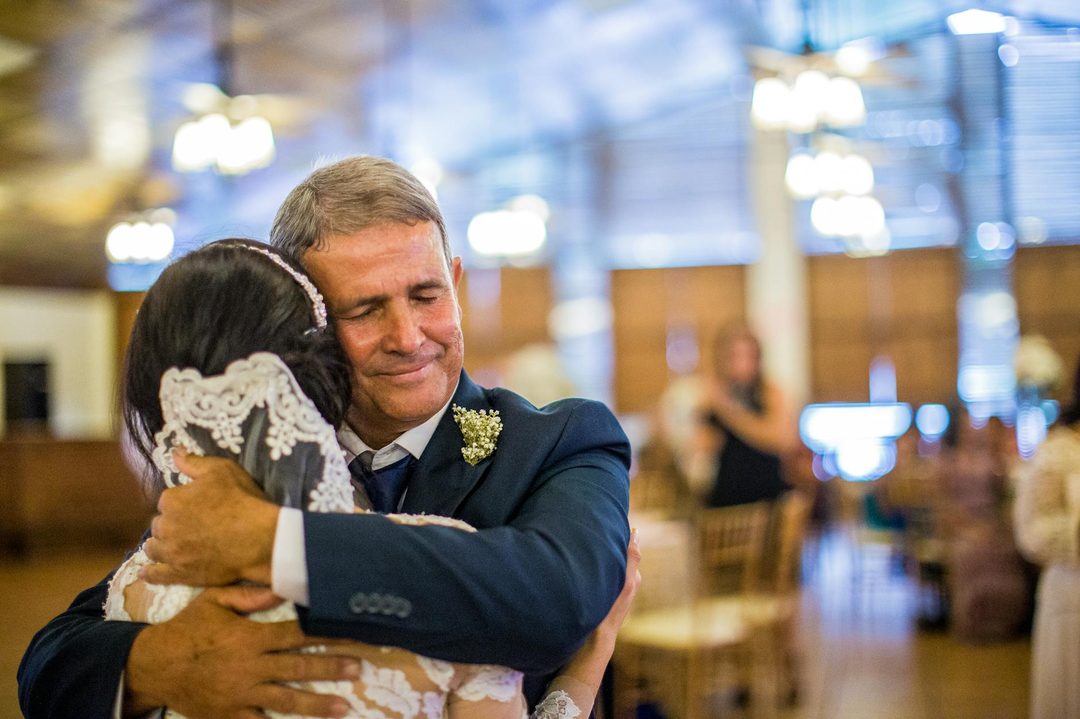 An older man in a navy suit and boutonniere embraces a woman wearing a white lace wedding dress and pearl headpiece at an elegant indoor reception venue with soft lighting and gues