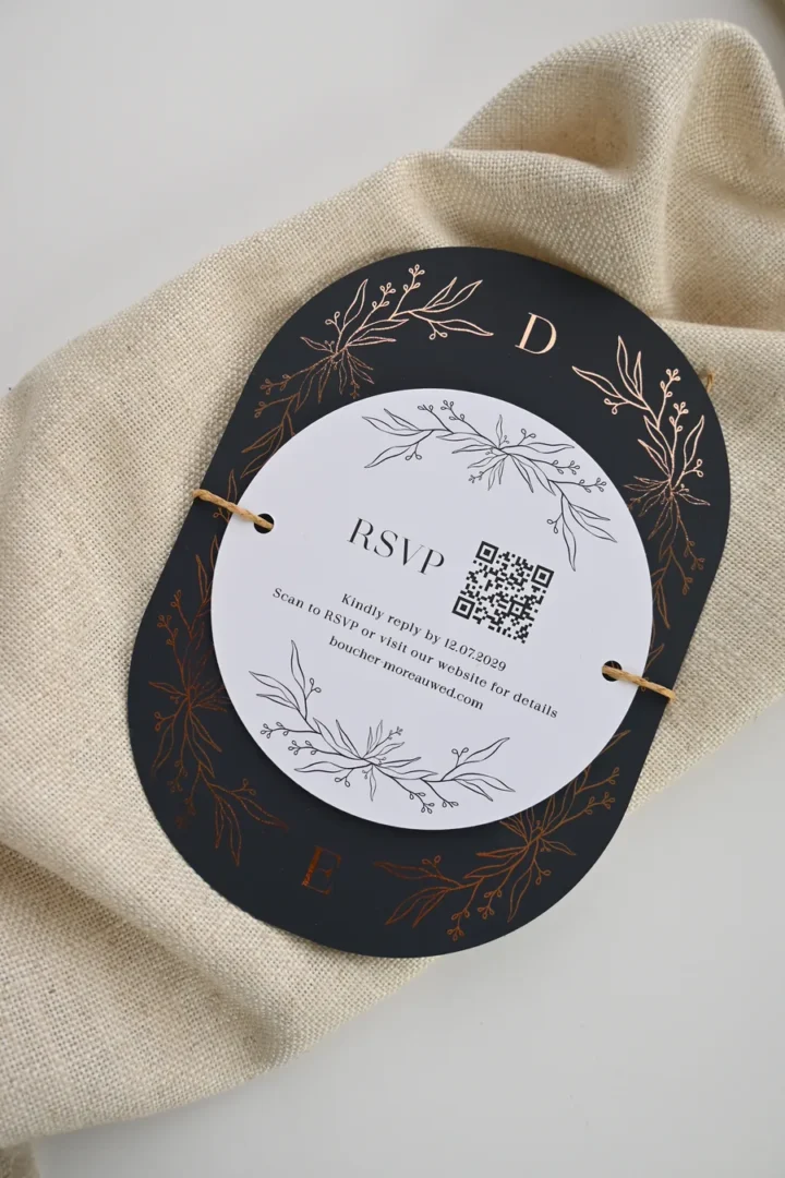 Paperlust Herbarium RSVP card, a white circle die-cut with botanical line art and QR code, tied with twine over a charcoal arch card with copper foil botanicals