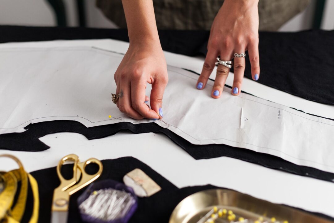 Close-up of a seamstress's hands using fine pins and tailor's chalk to mark a hem on a lace-edged wedding gown spread...