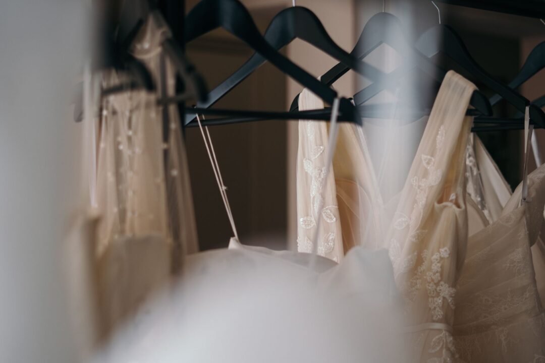 Three white and ivory wedding gowns hanging on a rack in a softly lit bridal boutique, with a seamstress's workbench in...