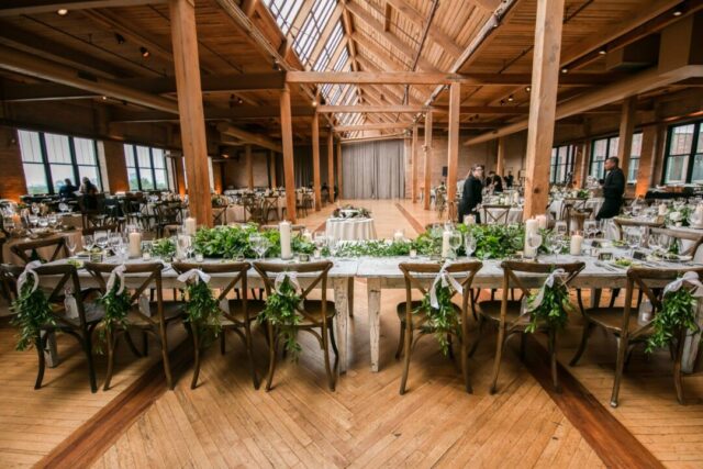 Long wooden farm table set for a wedding reception with small favor boxes tied in twine at each place setting, jars of...