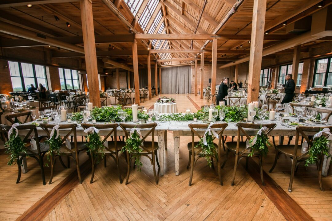 Long wooden farm table set for a wedding reception with small favor boxes tied in twine at each place setting, jars of...