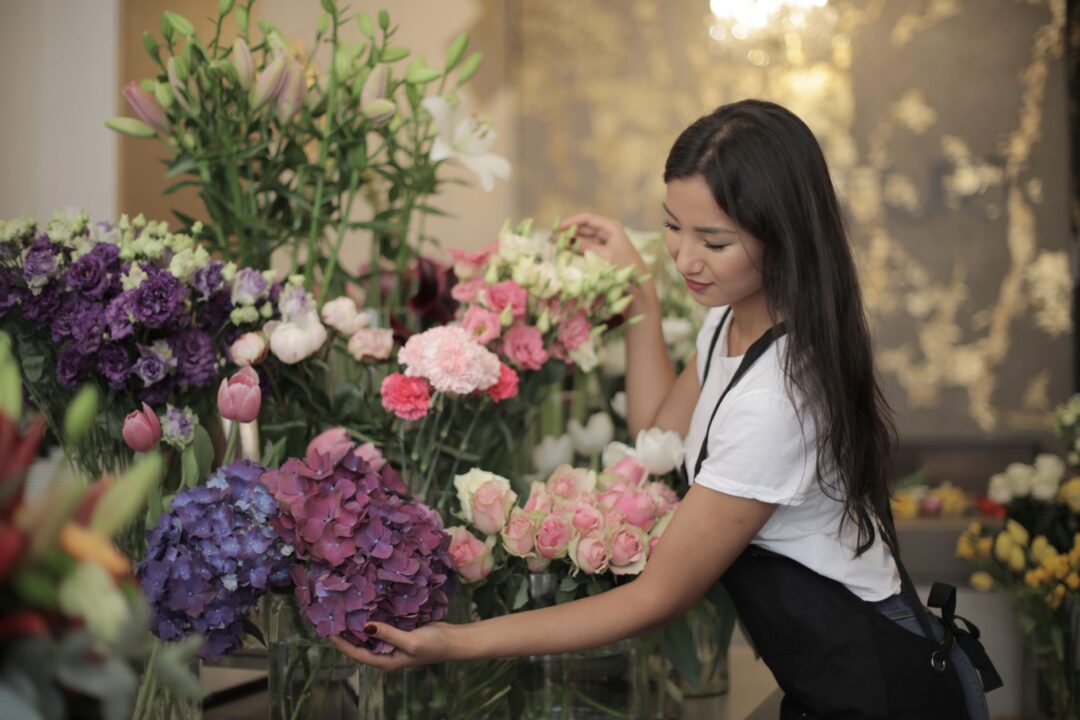 Professional florist in an apron arranging white hydrangeas and garden roses into a tall glass vase centerpiece at a...