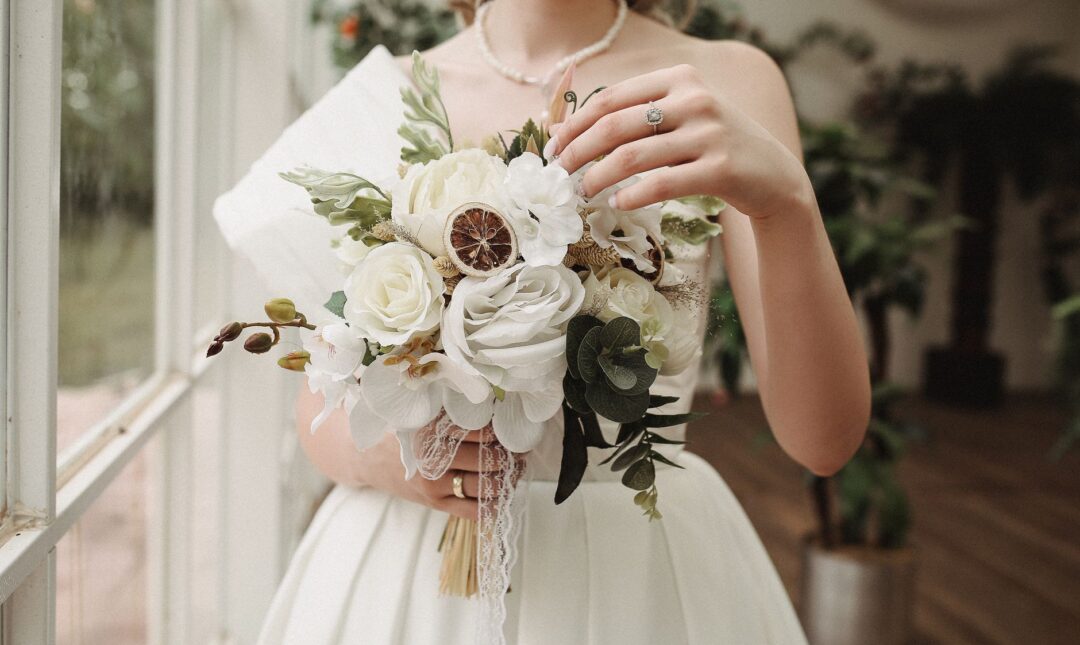 Bride holding a lush bridal bouquet of white peonies, garden roses, and trailing eucalyptus, photographed from above on a...