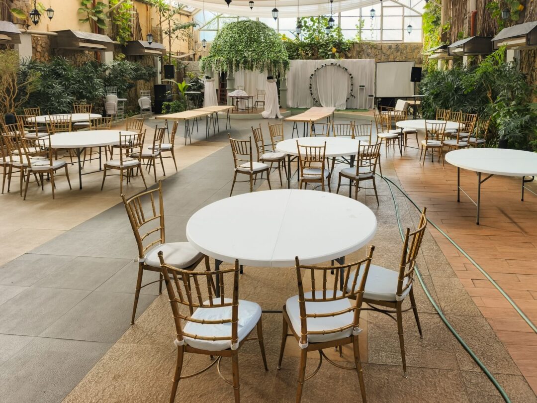 Close-up of chiavari chairs arranged around a round reception table set with white linens and a low floral centerpiece at...