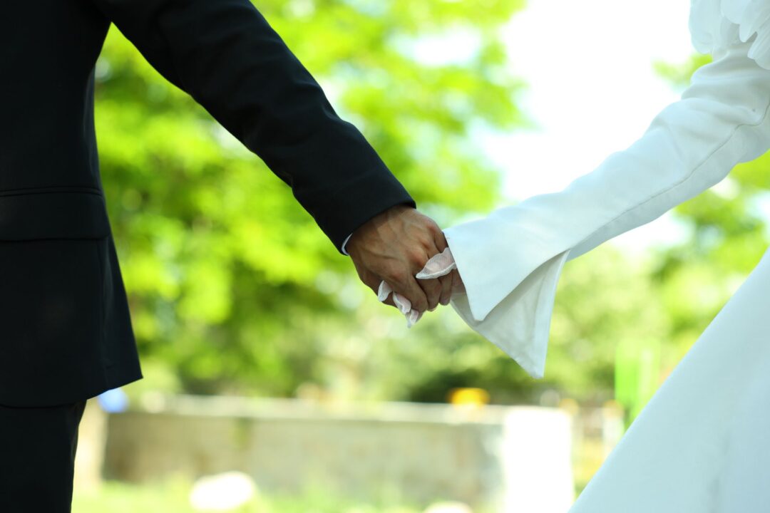 Bride and groom walking hand-in-hand through a white sailcloth tent at an outdoor summer wedding, elegant round tables...