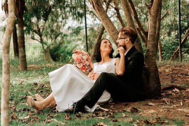 Couple in relaxed summer attire smiling under a wooden arch wrapped in white florals in a private backyard ceremony...