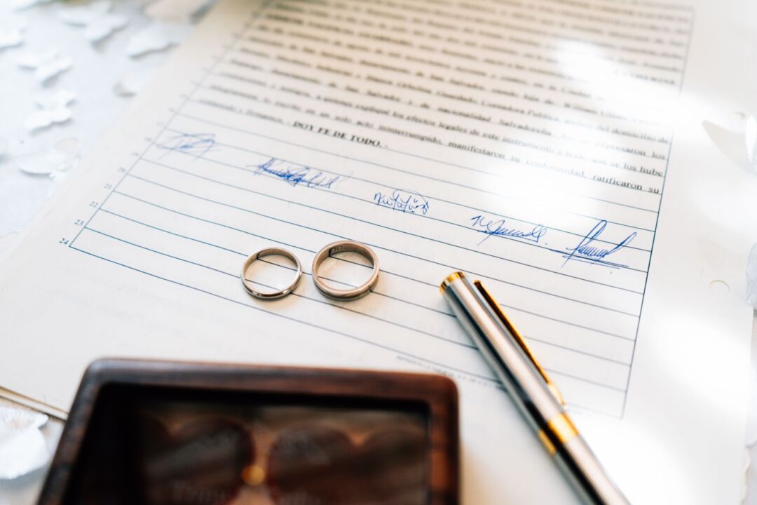 Marriage license and pen resting on a clerk's wooden desk with a small gold ring beside them, warm natural light